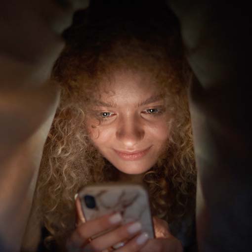 Woman checking phone at night under bedsheets.